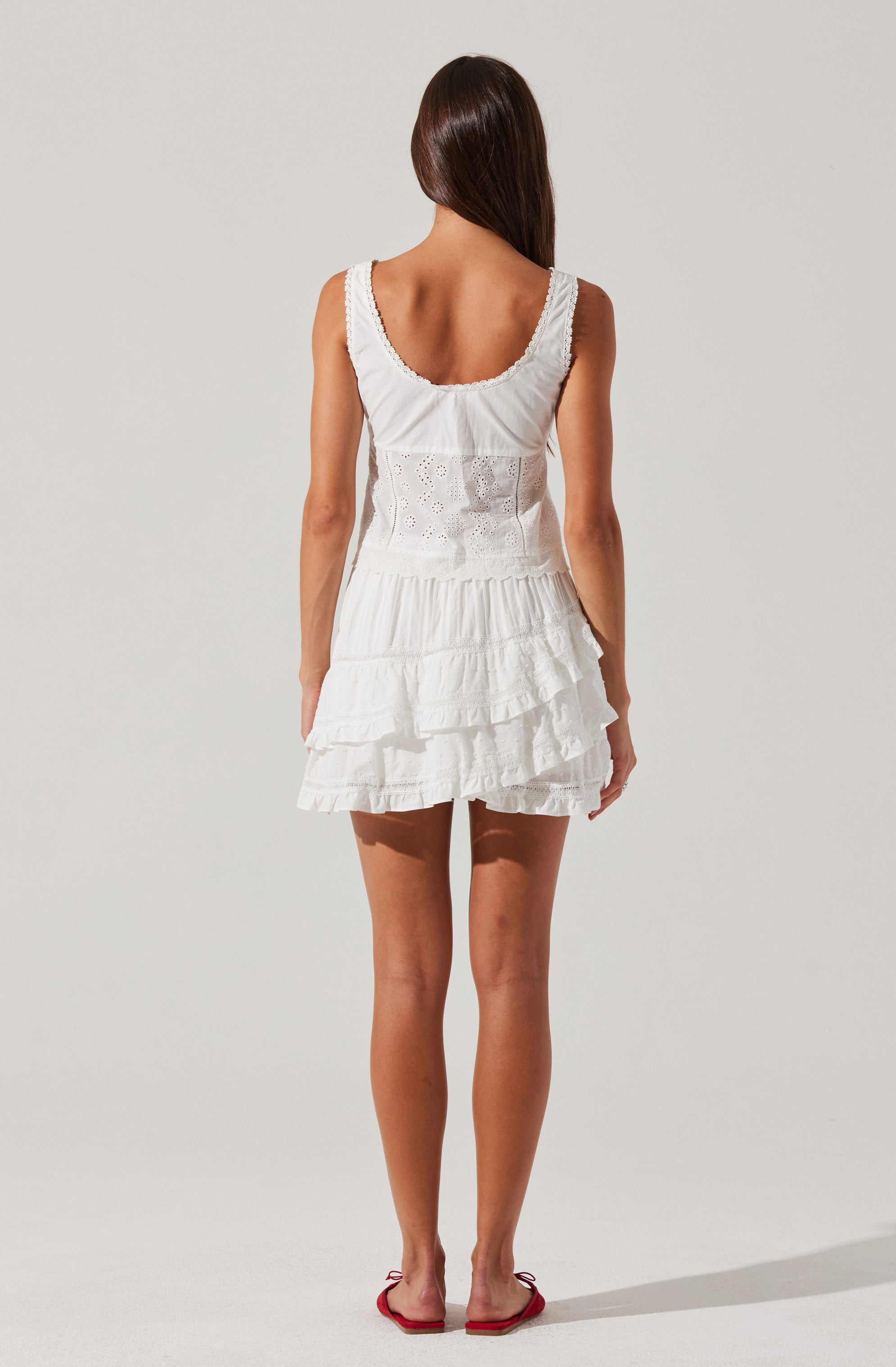 A woman is wearing a sleeveless white dress with a rounded neckline. The dress features embroidered detailing on the bodice and a tiered, ruffled skirt. She is also wearing flip-flops with a red strap.