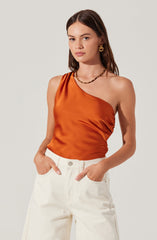 A woman is wearing a sleeveless, one-shoulder top made of shiny, burnt orange satin fabric. She has paired it with high-waisted, off-white pants. She accessorized with gold hoop earrings and a beaded necklace.