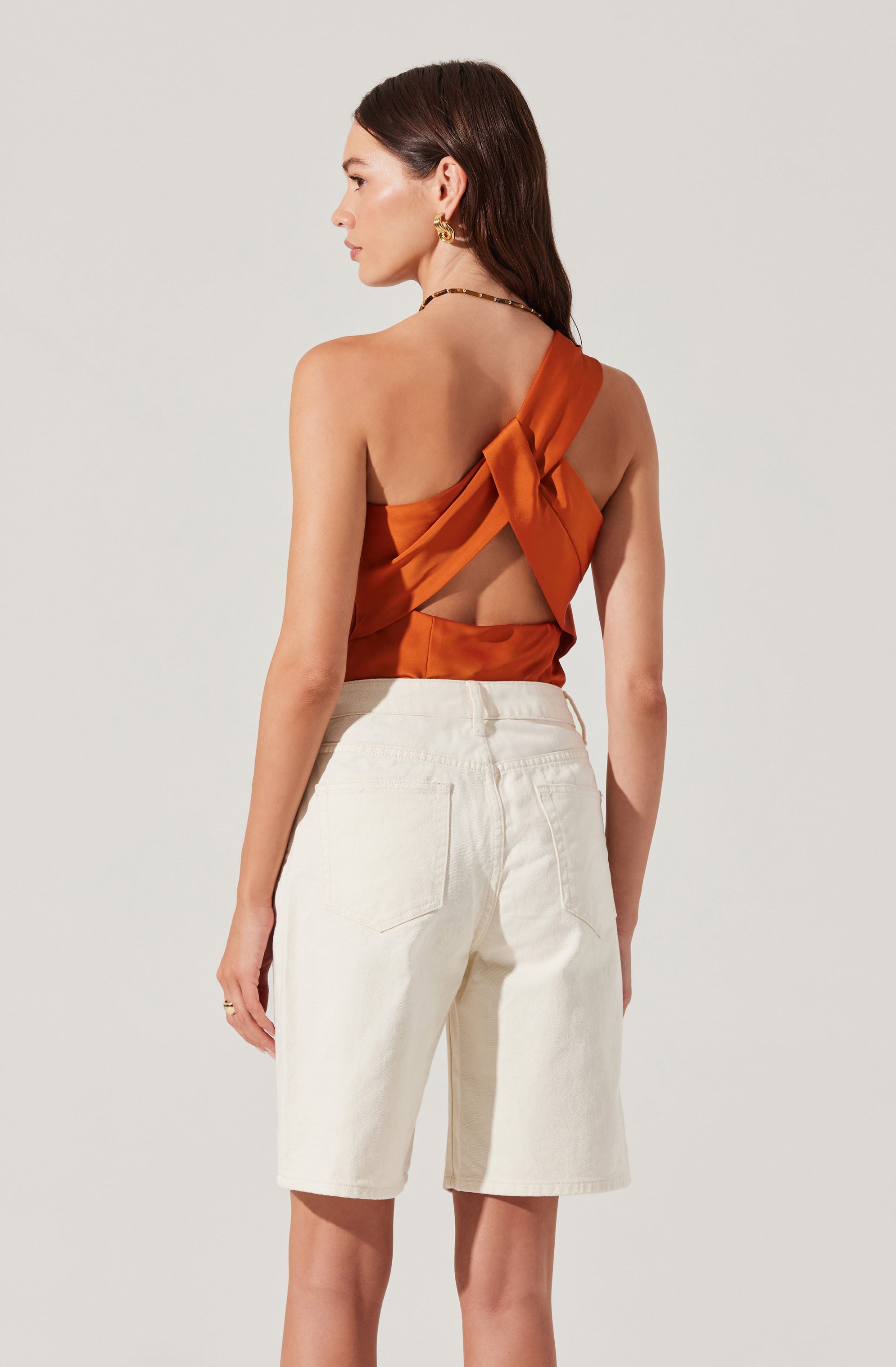A woman is wearing an orange satin sleeveless top with a large bow at the back and a cutout design, paired with high-waisted, knee-length white denim shorts. She has long dark hair, gold hoop earrings, and a black beaded necklace.