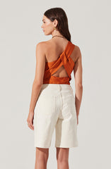 A woman is wearing an orange satin sleeveless top with a large bow at the back and a cutout design, paired with high-waisted, knee-length white denim shorts. She has long dark hair, gold hoop earrings, and a black beaded necklace.