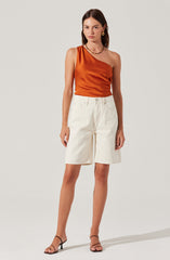 The woman is wearing a one-shoulder satin top in a rich, burnt orange color. She pairs it with high-waisted, loose-fitting white shorts. She is also wearing black strappy high-heeled sandals and accessorizing with gold hoop earrings and a chain necklace.