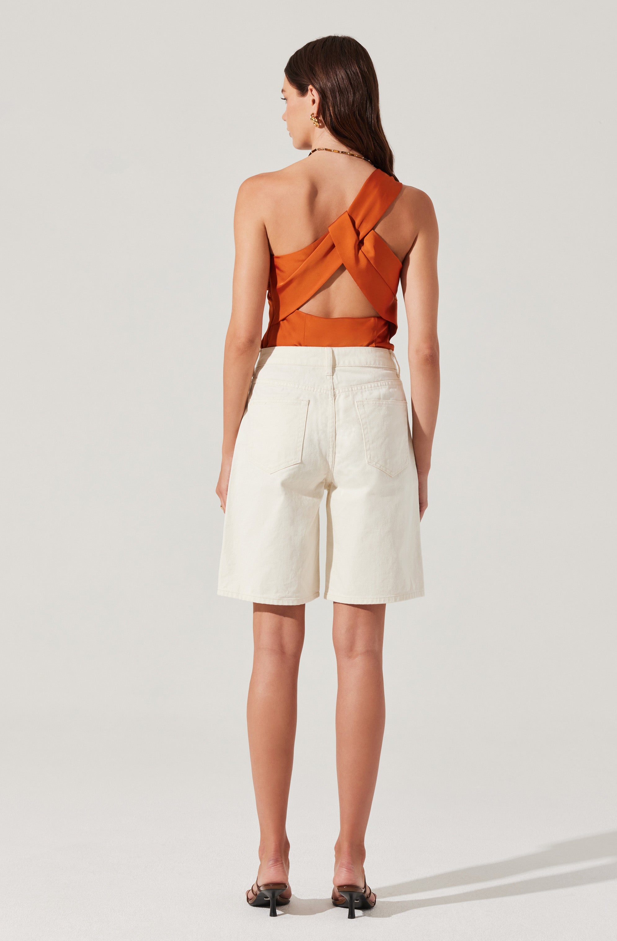 A woman is wearing a sleeveless, rust-colored satin top with a knotted and crisscross design at the back, revealing an open-back style. She pairs it with high-waisted, light-colored knee-length shorts.