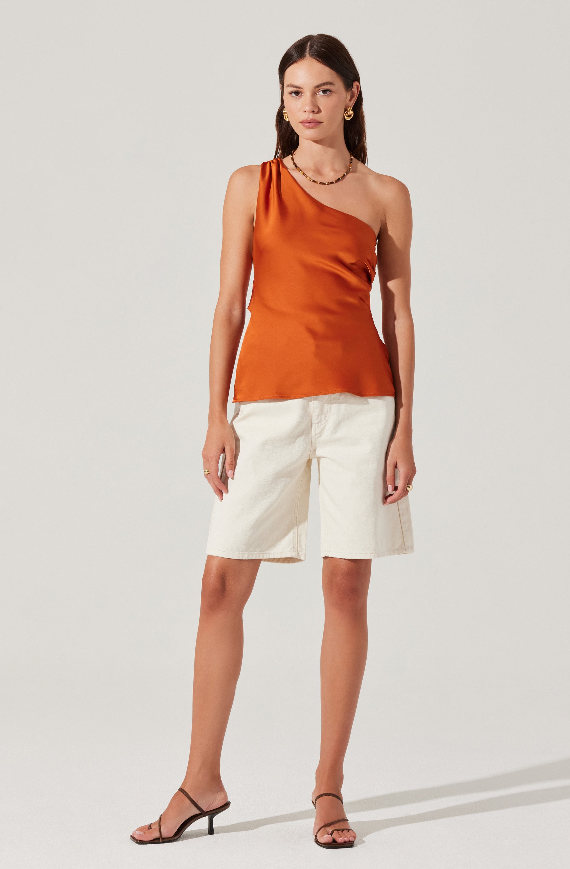 A woman is wearing an orange satin one-shoulder top with a draped design on the left shoulder, paired with high-waisted, knee-length white shorts. She is also wearing black strappy high-heeled sandals, gold hoop earrings, and a gold necklace.
