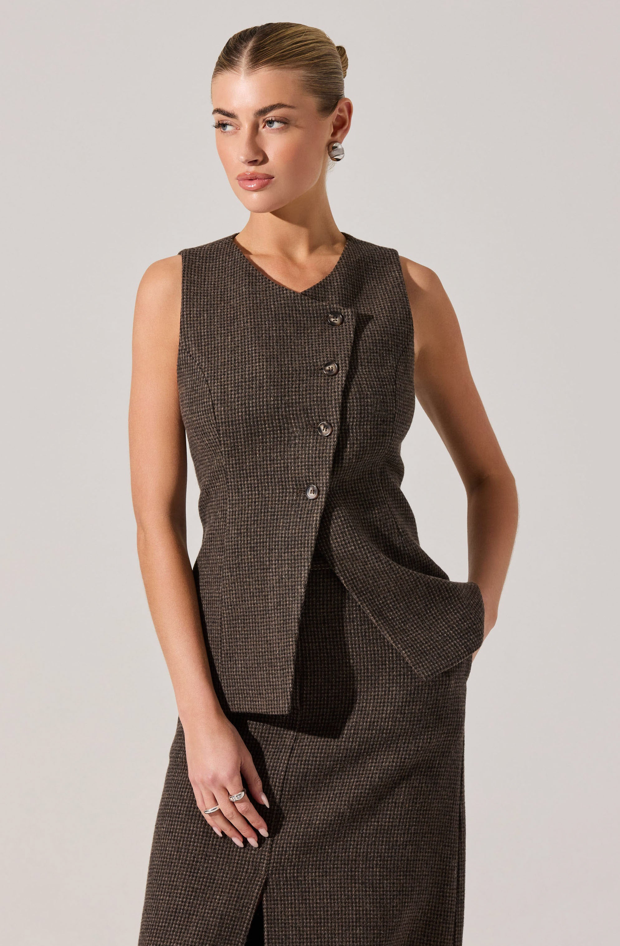 A woman is wearing a sleeveless, knee-length skirted suit in a dark, checkered pattern. The suit has a tailored top with an asymmetrical button closure and a matching skirt with a front slit. She has her left hand in her skirt pocket and is accessorized with earrings and rings.