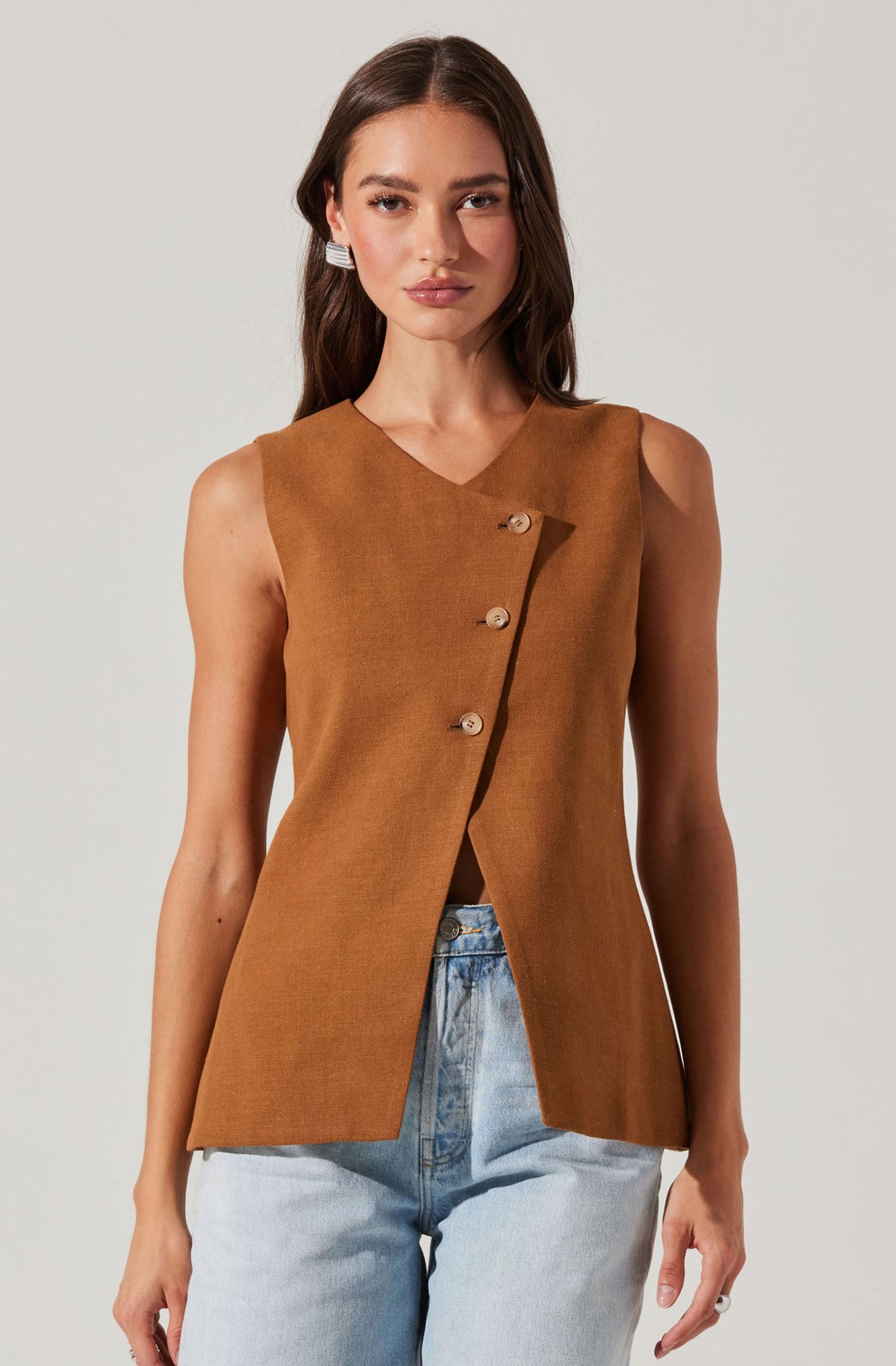 A woman is wearing a sleeveless, camel-colored vest with a V-neckline. The vest has three large, round buttons running diagonally across the front. It has a tailored fit and extends slightly below the waist. She pairs it with light blue, high-waisted jeans.