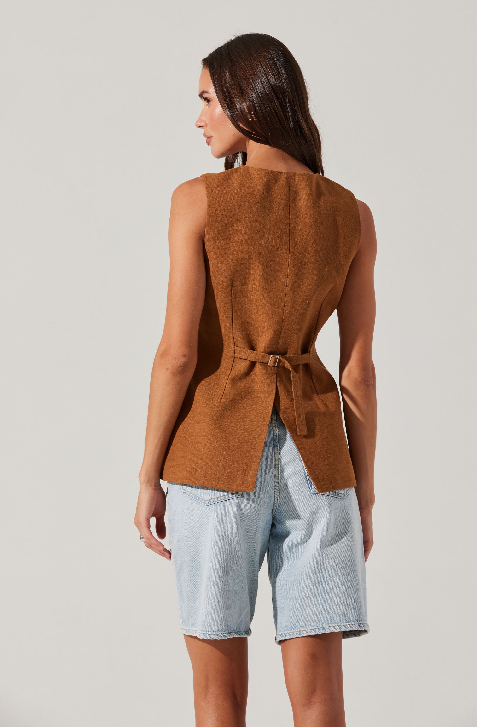 A woman is wearing a sleeveless, camel-colored top with a belt that ties at the waist and a slit detail at the back. She pairs it with light blue denim shorts. The top has a smooth texture and a structured design.