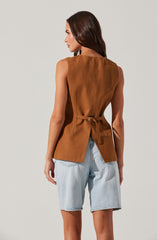 A woman is wearing a sleeveless, camel-colored top with a belt that ties at the waist and a slit detail at the back. She pairs it with light blue denim shorts. The top has a smooth texture and a structured design.