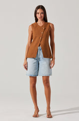 The woman is wearing a sleeveless, tailored brown vest with a structured fit and four buttons down the front. She pairs it with light-wash denim shorts that are knee-length and have a relaxed fit. She completes the outfit with nude high-heeled sandals.