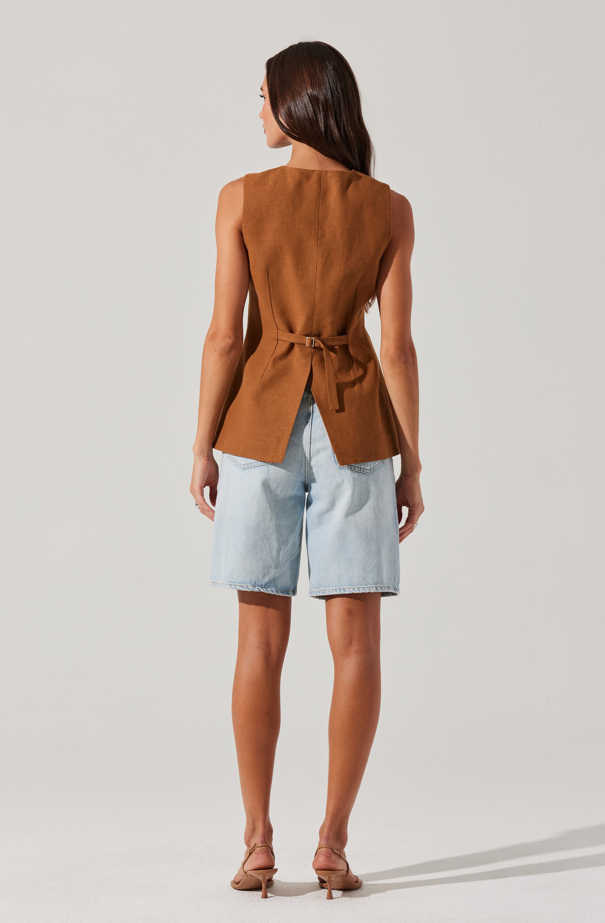 The image shows a woman wearing a sleeveless, camel-colored vest with a tied waist detail. The vest has a small slit at the back hem. She pairs it with light blue denim knee-length shorts. She is also wearing beige high-heeled sandals.