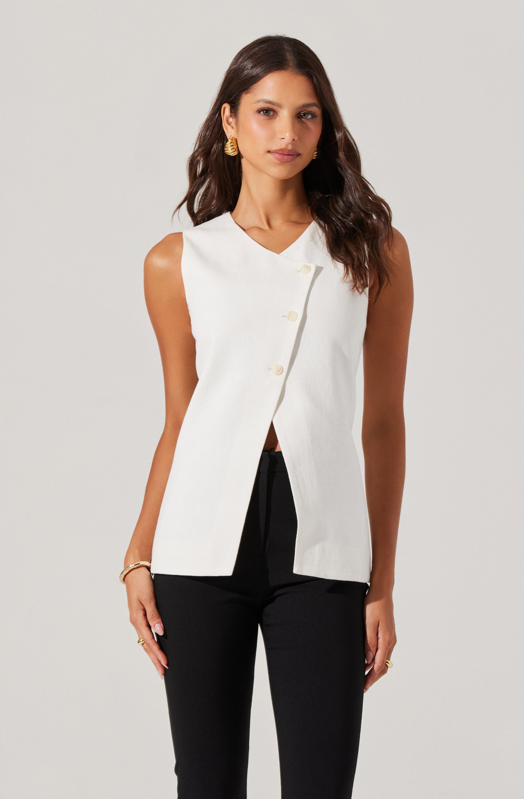 A woman is wearing a sleeveless, white vest with a V-neckline and the vest is fastened with large cream-colored buttons down the front. She pairs it with black pants.