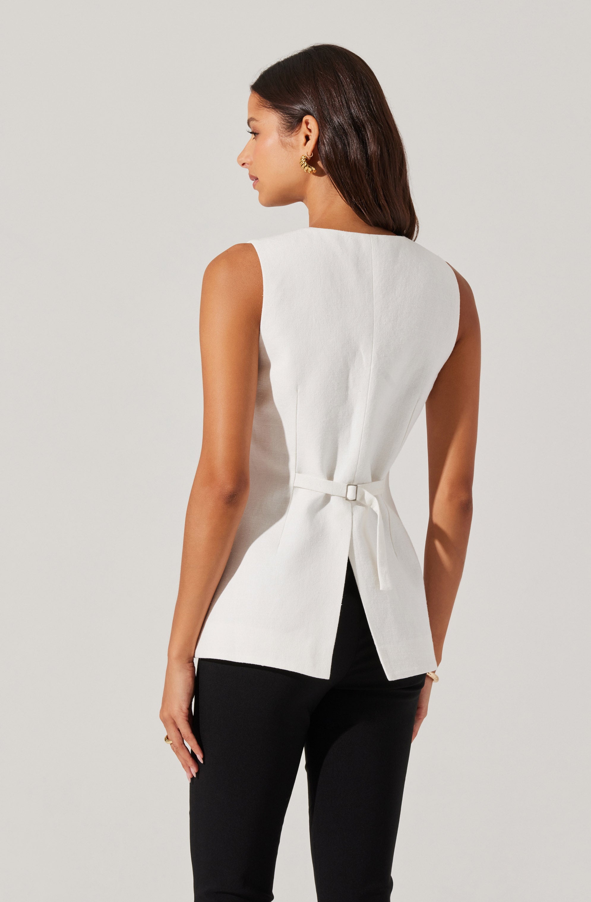 A woman is wearing a sleeveless, white, tailored top with a boat neckline and a waist tie, paired with black pants. The top has a structured fit with a back slit. She has dark brown hair and is accessorized with gold hoop earrings.
