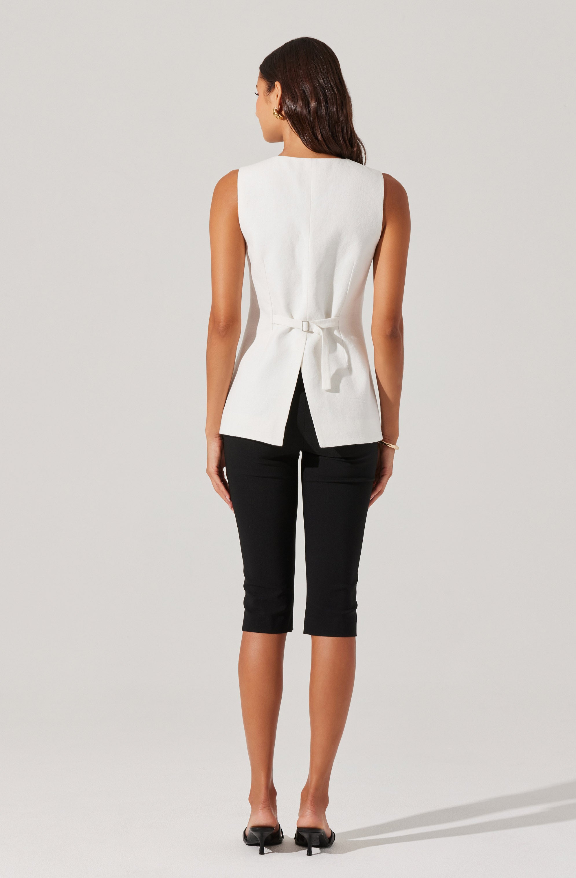 A woman is wearing a sleeveless, white, tailored top with a back tie detail, paired with black, knee-length fitted pants. She has dark, shoulder-length hair and is wearing small earrings and a bracelet on her left wrist. She is standing against a plain, light-colored background in a relaxed pose.
