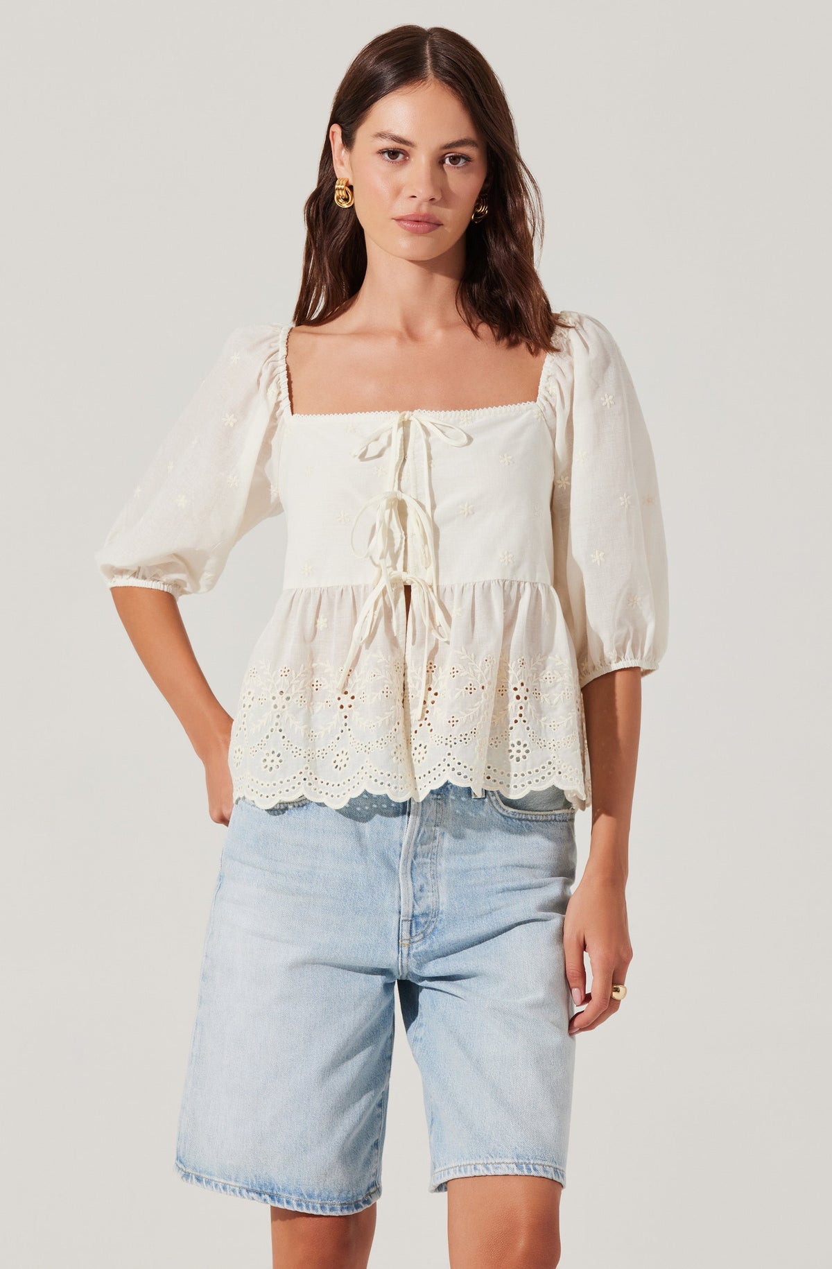 A woman is wearing an off-white, square-neck blouse with puffed three-quarter sleeves. The blouse features decorative eyelet embroidery and a ruffled hem, with ties at the front for adjustment. She is pairing it with light-wash denim shorts.