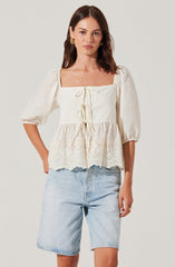 A woman is wearing an off-white, square-neck blouse with puffed three-quarter sleeves. The blouse features decorative eyelet embroidery and a ruffled hem, with ties at the front for adjustment. She is pairing it with light-wash denim shorts.