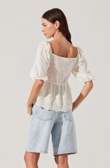 A woman is wearing an off-white, puff-sleeve blouse with embroidered floral details and eyelet lace trim at the hem. She pairs it with light-wash, knee-length denim shorts. She has long, dark brown hair and is accessorized with gold hoop earrings.