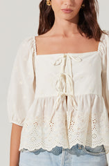 A woman is wearing an off-white, square-neck blouse with puffed three-quarter sleeves. The blouse features embroidered floral patterns and has a front tie detail with three bows, along with a scalloped hem with intricate eyelet embroidery.