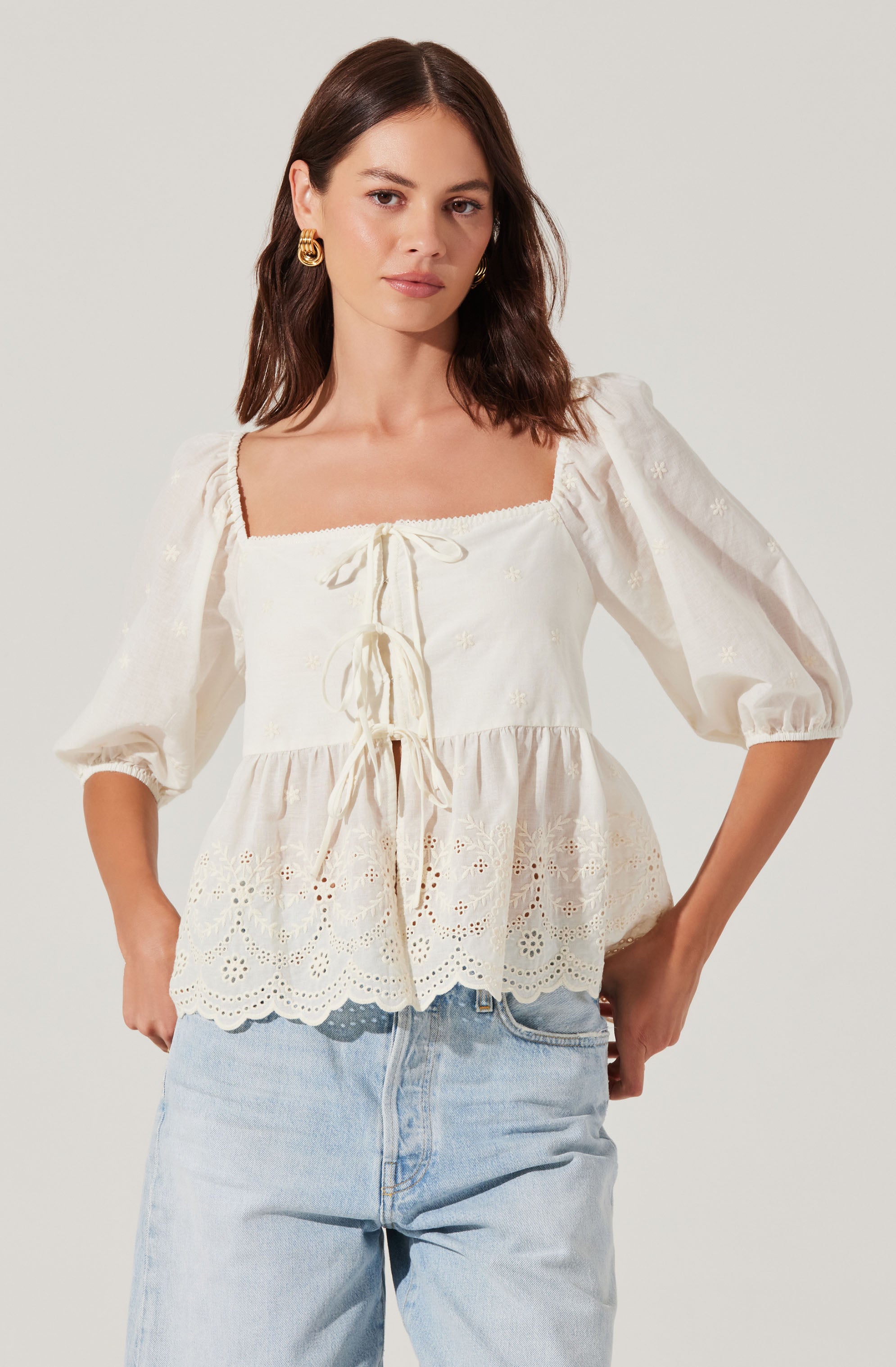 A woman is wearing an off-white, square-neck blouse with puffed three-quarter length sleeves. The blouse features delicate embroidery and eyelet lace details, with a tied drawstring at the front. The hem has a scalloped edge with intricate embroidery. She pairs the blouse with light-wash jeans and gold hoop earrings.