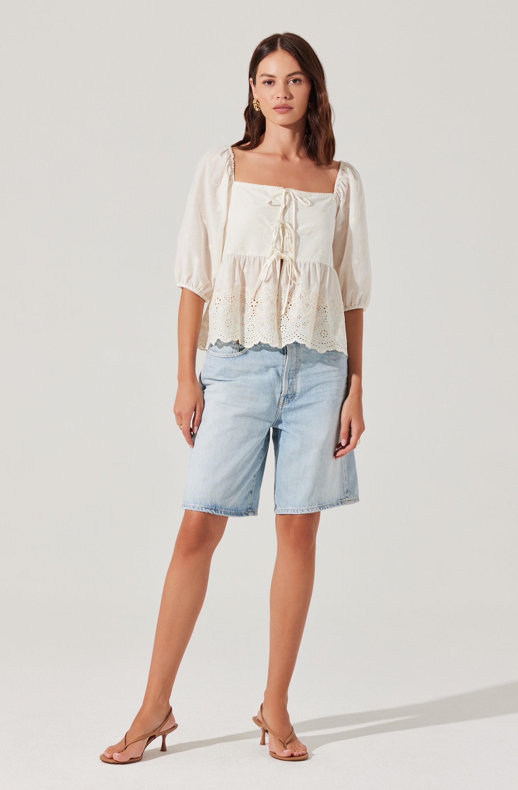 A woman is wearing a cream-colored, square-neck blouse with puffed three-quarter sleeves. The blouse has a front tie detail and a lace or eyelet pattern at the hem. She is paired with light-wash denim knee-length shorts and nude-colored, open-toe high-heeled sandals.