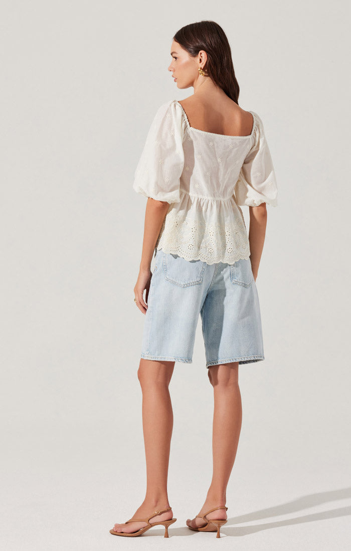 A woman is wearing a white blouse with puffed sleeves and eyelet embroidery details, featuring a gathered waist and a scalloped hem. She pairs it with light-wash denim knee-length shorts and beige open-toe heeled sandals.