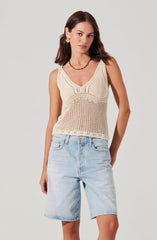 A woman wearing a sleeveless, cream-colored, crocheted tank top with a V-neckline and intricate pattern design, paired with light-wash denim Bermuda shorts. She accessorizes with gold earrings and a beaded necklace.
