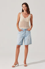 A woman is wearing a sleeveless, cream-colored, crocheted knit top with a V-neckline and intricate open-knit patterns. She has paired it with light-wash, knee-length denim shorts and nude open-toe high-heeled sandals.