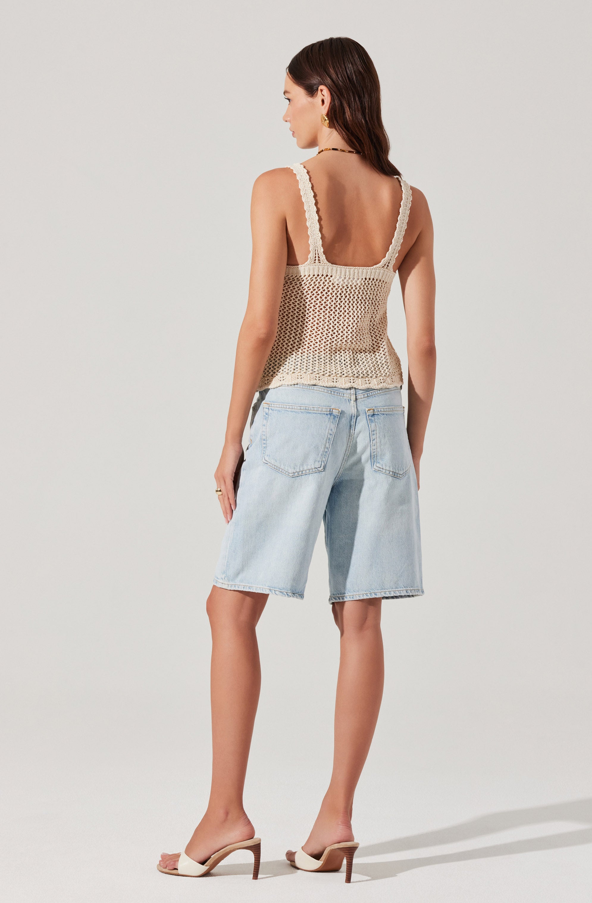 A woman is wearing a sleeveless, cream-colored crochet top with thick straps and a loose fit. She pairs it with light-wash denim shorts that have a relaxed fit and large back pockets. She completes her outfit with white, open-toed high-heeled sandals featuring a single strap across the toes and a slim heel.
