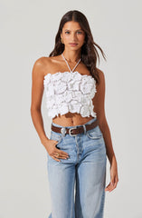 A young woman is wearing a white, strapless, crocheted crop top with a textured, ruffled design. She has accessorized with gold hoop earrings and a thin necklace. She is also wearing light blue, high-waisted jeans with a brown belt.