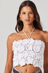 A woman is wearing a sleeveless, strapless white top made of textured fabric with large, ruffled floral appliqués and open-knit details. She accessorizes with gold hoop earrings and a white, knitted tie around her neck.