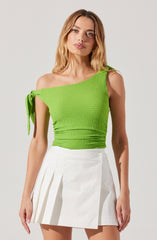 A woman is wearing a bright green, textured, sleeveless top with a one-shoulder design. The top features a tied shoulder strap on her left side. She pairs it with a high-waisted, white mini skirt that has pleats on the left side.