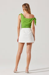 The image shows a woman dressed in a bright green, textured top with ruffled, tied shoulder straps, paired with a white, high-waisted mini skirt. She is wearing white high-heeled shoes and has long, straight hair.