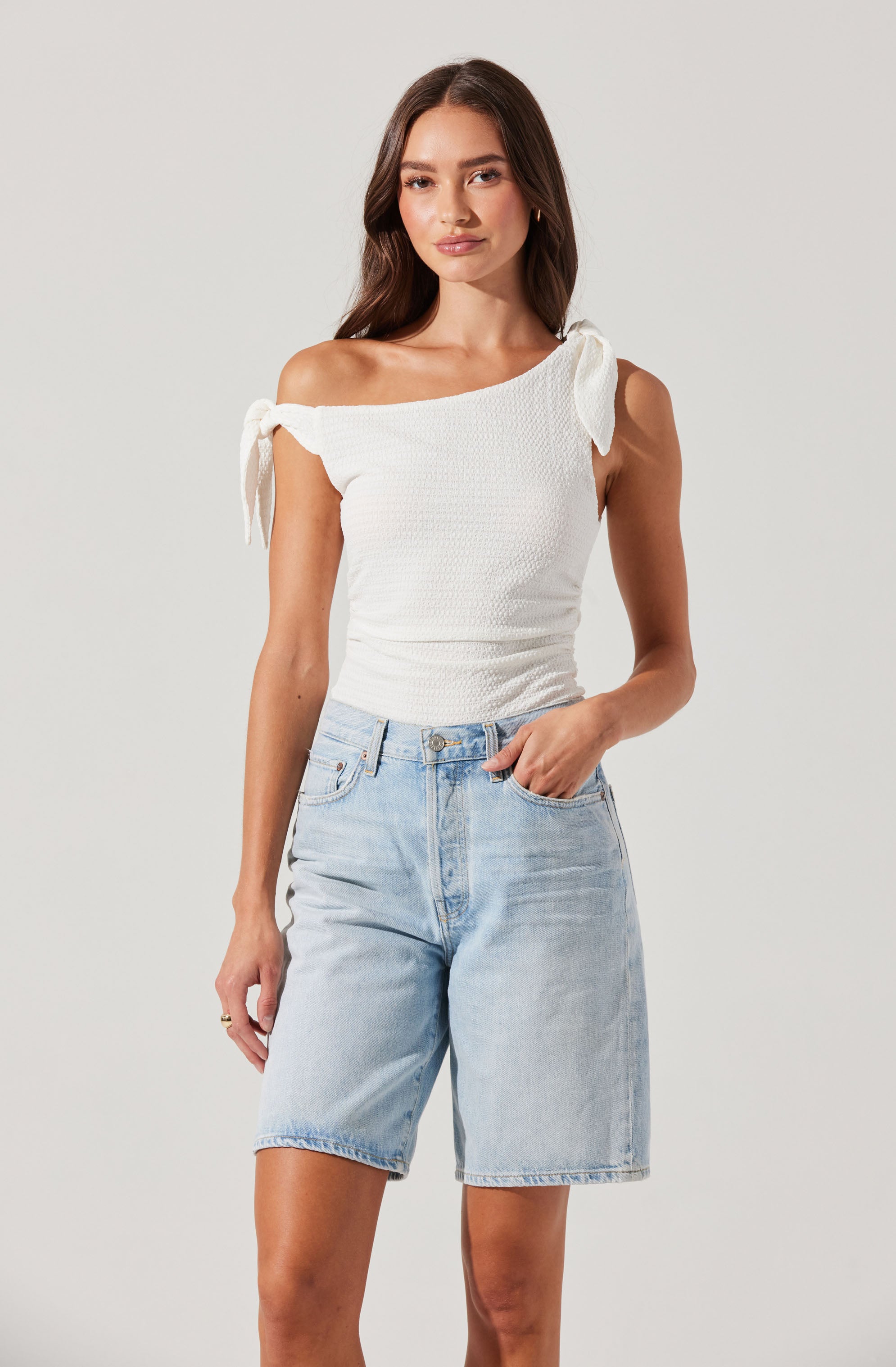 A woman is wearing an off-the-shoulder white top with textured fabric and tied shoulder straps, paired with high-waisted, light-wash denim shorts.