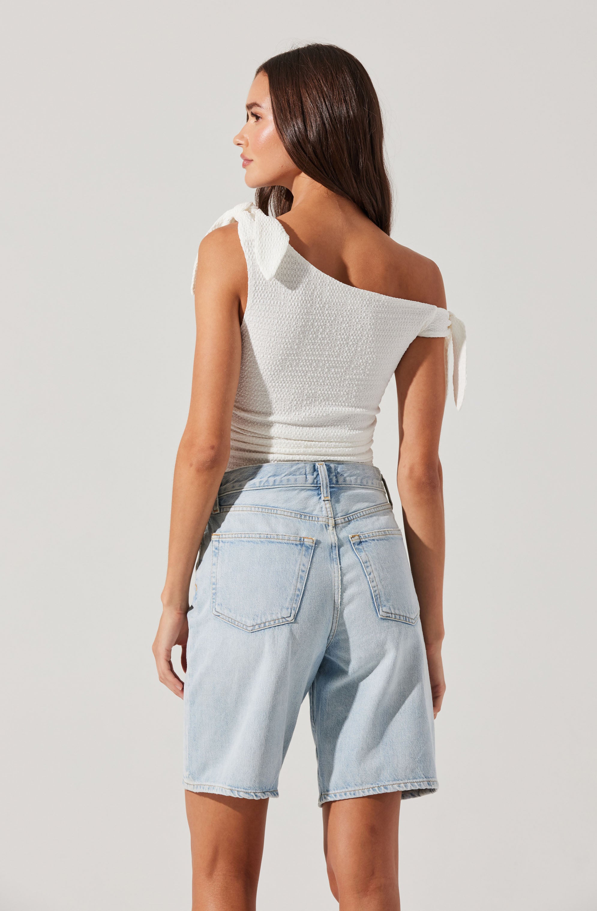 A woman is wearing a white sleeveless top with textured fabric and shoulder ties, paired with light wash denim Bermuda shorts.