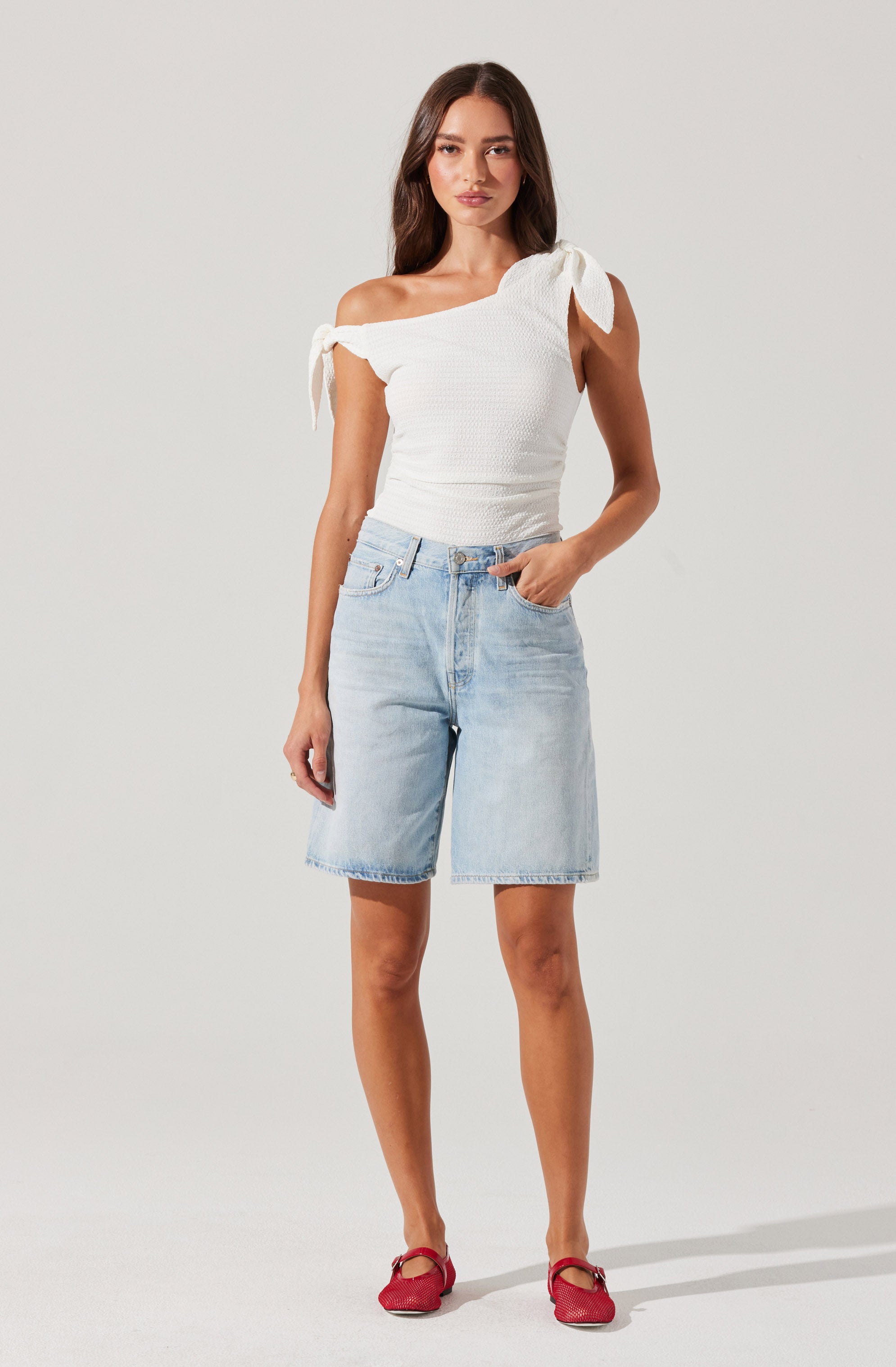 A woman is wearing a white, textured, one-shoulder top with shoulder ties and bow details on each shoulder. She is also wearing light-wash, knee-length denim shorts with front pockets. She has on red shoes with ankle straps.