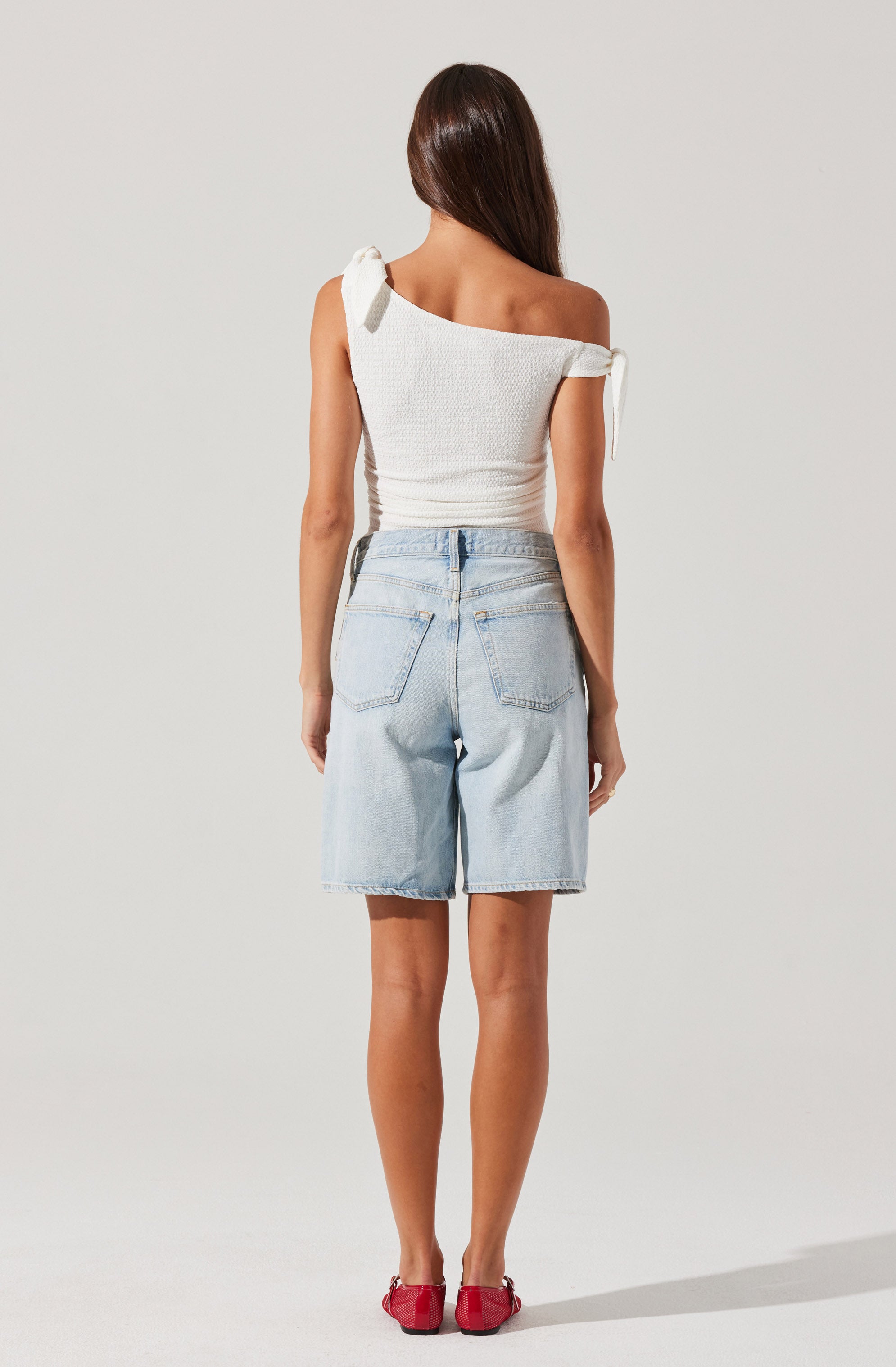 A woman is wearing a white, textured, off-the-shoulder top with tied shoulder straps. She is also wearing light blue, knee-length denim shorts with back pockets, and red shoes.