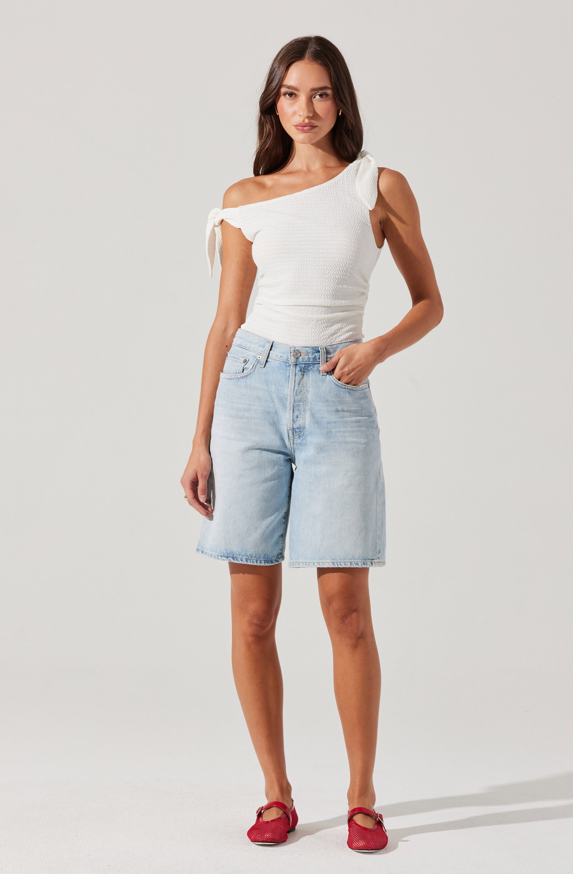 A woman is wearing a white, sleeveless, one-shoulder top with a textured pattern and tied shoulder straps. She pairs it with light-wash, knee-length denim shorts. She completes the outfit with red flat shoes featuring a bow detail.