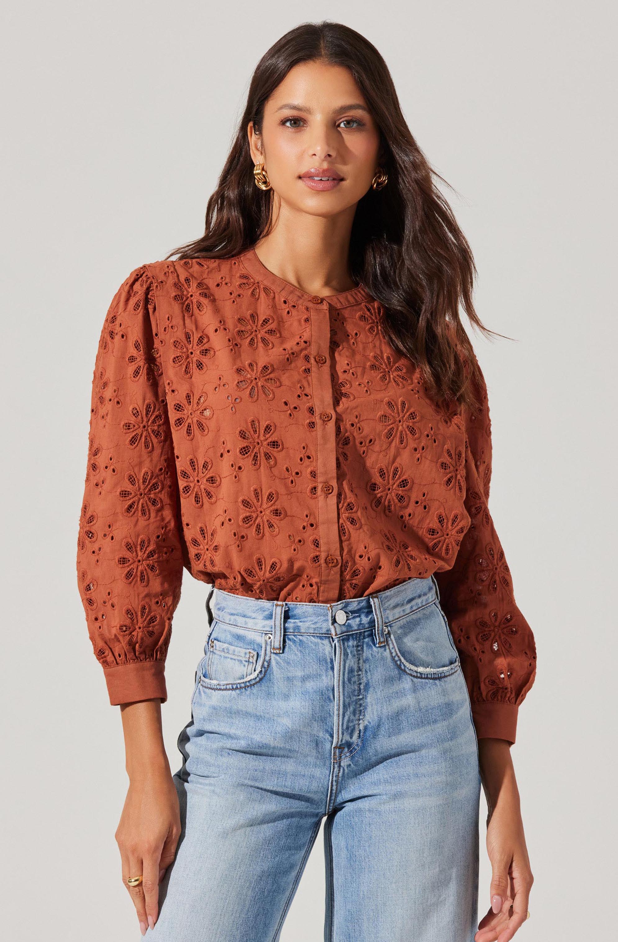 A woman is wearing a long-sleeved, rust-colored blouse with eyelet floral embroidery and puffed shoulders. The blouse has a round neckline and buttons down the front. She pairs it with high-waisted, light-wash jeans.