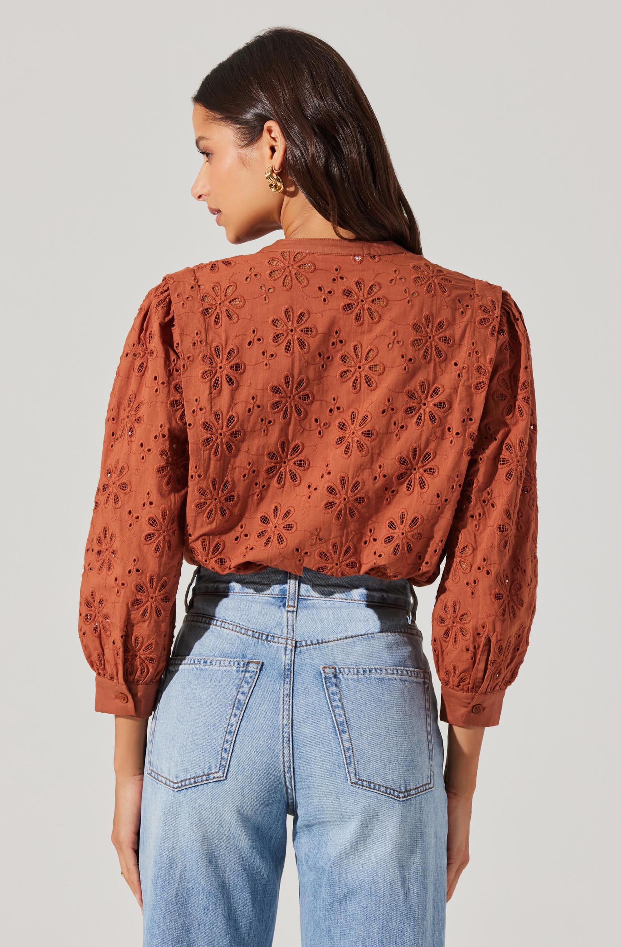 A woman is wearing a rust-colored, long-sleeve blouse with an eyelet floral pattern, featuring puffed sleeves gathered at the cuffs. She has paired the blouse with high-waisted light blue jeans.