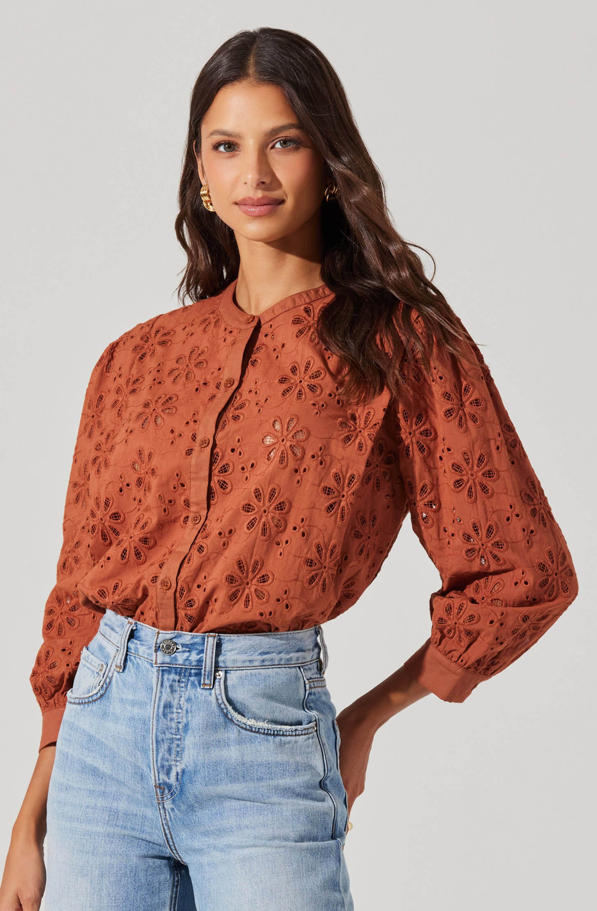A woman is wearing a rust-colored, long-sleeved blouse with an eyelet floral pattern. The blouse has a front button closure and a round neckline. The sleeves are gathered at the cuffs, giving a slightly puffed appearance.