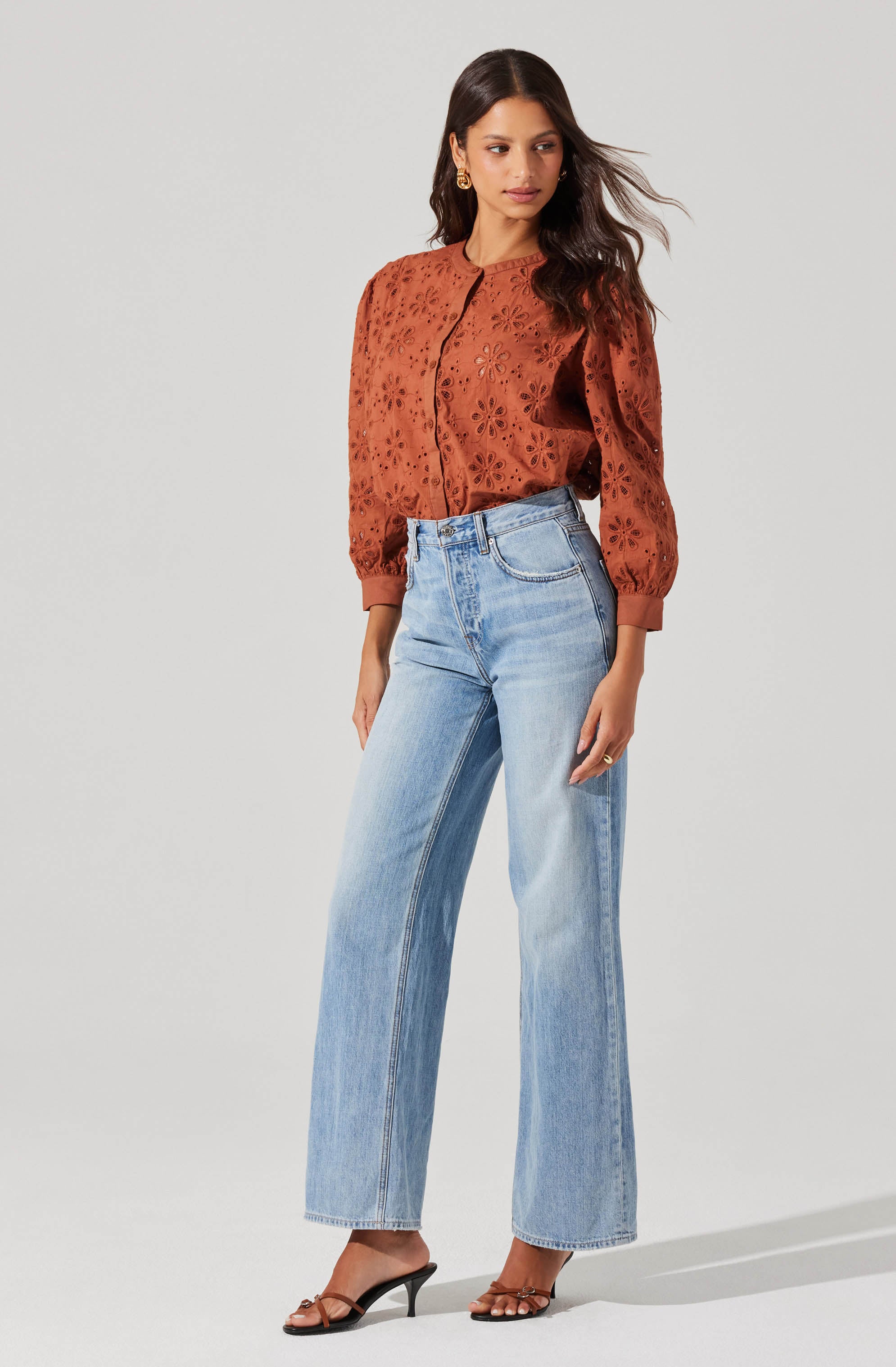 A woman is wearing a rust-colored, long-sleeved blouse with eyelet embroidery and a round neckline. The blouse has a button-down front and the sleeves are gathered at the cuffs. She is also wearing light blue, high-waisted, wide-leg jeans with a classic five-pocket design. She has on black, open-toe high-heeled sandals with thin straps.