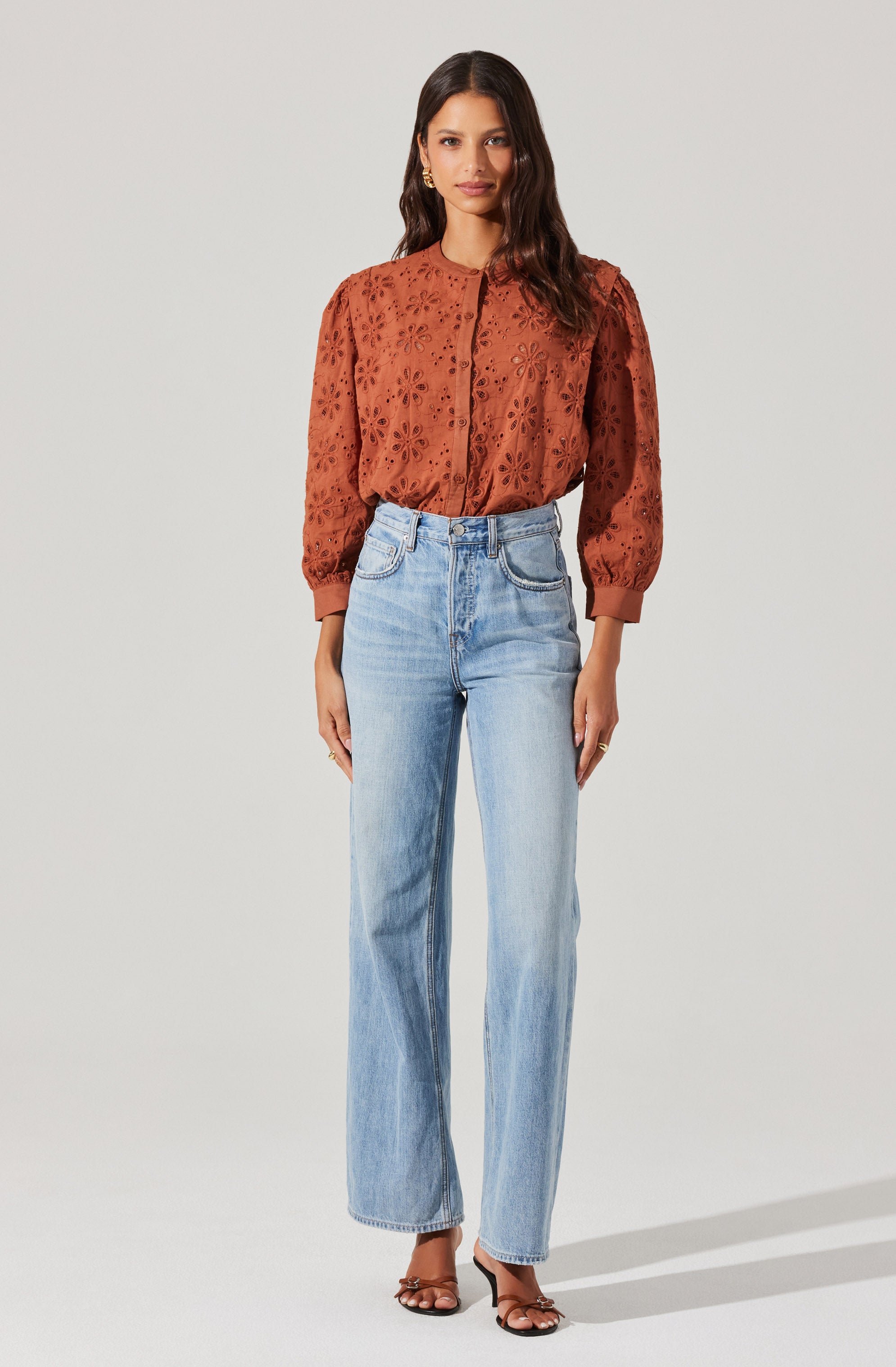 A woman is wearing a burnt orange blouse with embroidered floral cutouts and puffed long sleeves that gather at the cuffs. She pairs it with high-waisted, light-wash straight-leg jeans and black strappy heels.