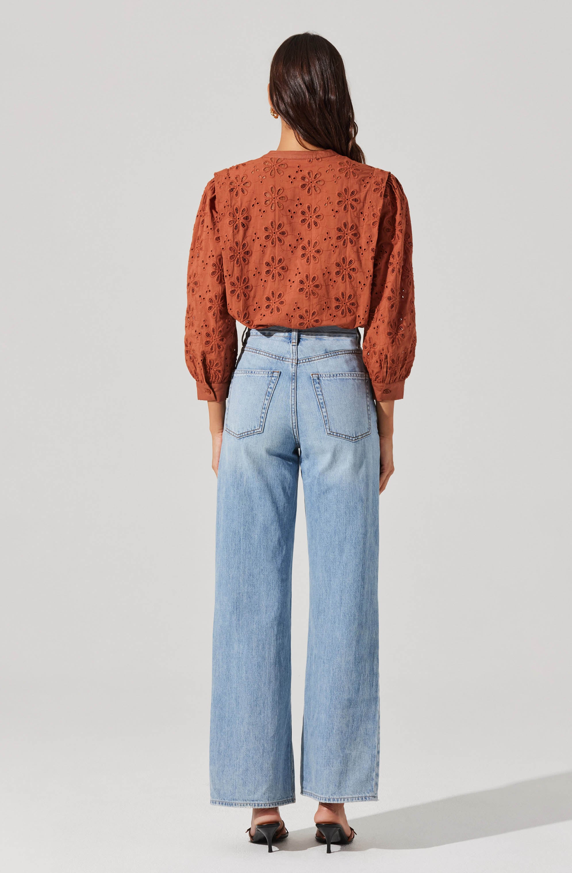 A woman is wearing a rust-colored blouse with embroidered floral cutouts and puffed long sleeves, paired with high-waisted, light-wash wide-leg jeans. She is also wearing black high-heeled shoes.