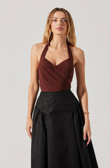 A woman is wearing a sleeveless, deep V-neck halter top in a dark burgundy color, featuring gathered fabric details across the bust. She is also dressed in a high-waisted, black skirt with a full, pleated design.