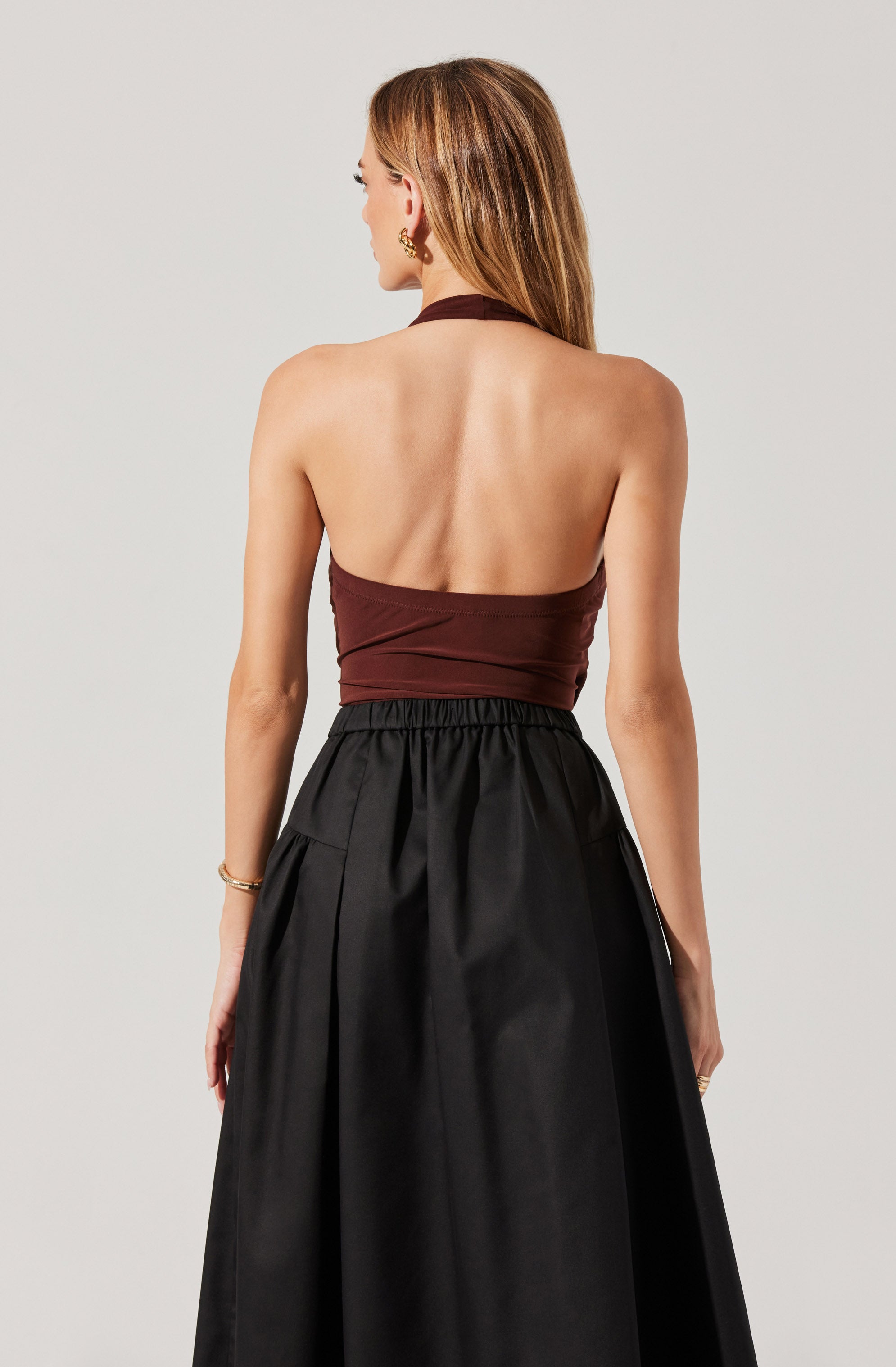 A woman is wearing a sleeveless halter top in dark brown with a gathered detail around the waist, paired with a high-waisted, full-length black skirt that has an elastic waistband and subtle pleats. She has long, straight hair and is accessorized with a gold earring and bracelet.