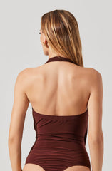 The image shows a woman with light brown, shoulder-length hair wearing a sleeveless, halter-style bodysuit in a deep chocolate brown color. The bodysuit has a high neckline and is fitted, accentuating her figure. The back of the bodysuit is open, exposing her shoulder blades and upper back.