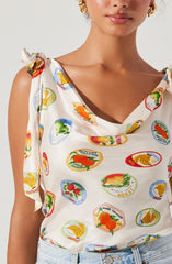 A woman is wearing a sleeveless, white top with colorful fruit-themed print patches, including oranges, strawberries, and bananas. The top has a draped neckline and tied shoulder straps with a bow detail. She is also wearing gold hoop earrings.