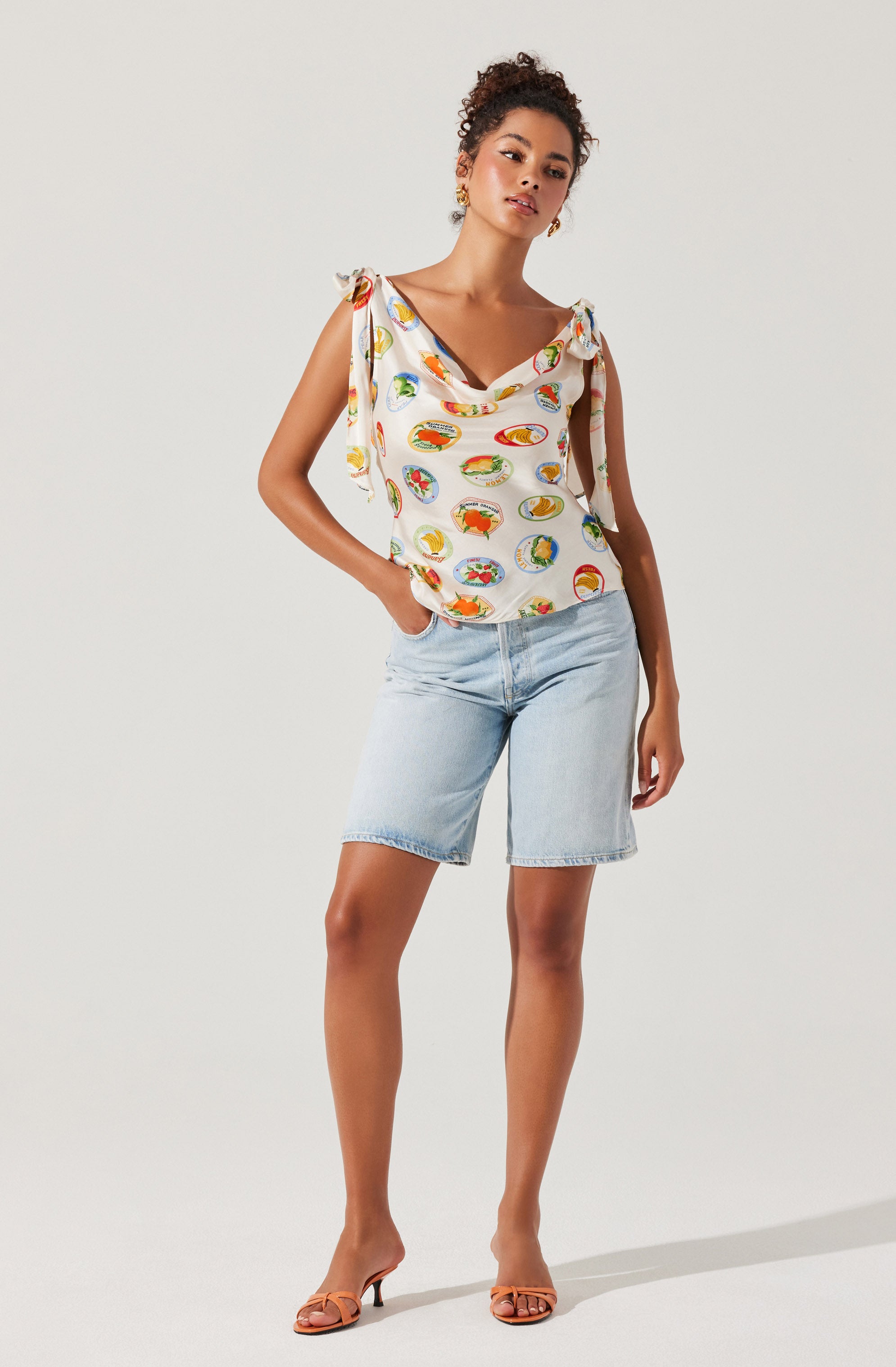 A woman is wearing a sleeveless, cream-colored top with tied shoulder straps and a colorful, circular pattern. She pairs it with light wash denim shorts and tan, open-toe heels.