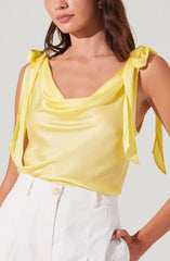 A woman is wearing a sleeveless yellow satin top with tie shoulder straps that form bows, and high-waisted white trousers with a button closure.