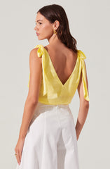 A woman is wearing a sleeveless, yellow top with shoulder ties that form bows, and white high-waisted, wide-leg pants. The top has a deep V-back, and she is accessorized with gold hoop earrings.