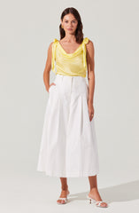 A woman is wearing a sleeveless yellow top with shoulder ties and a draped neckline. She is paired with high-waisted, wide-leg white pants that have pleats and large front pockets. She completes the outfit with white heeled sandals with thin straps.
