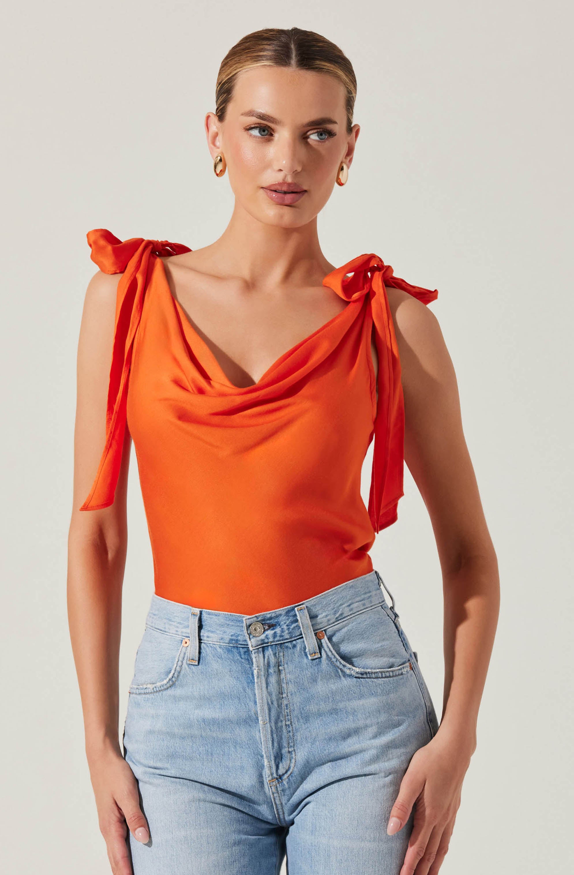 A woman is wearing a sleeveless, bright orange top with gathered fabric around the neckline and shoulder ties that form bows. She has paired it with high-waisted, light-wash jeans.